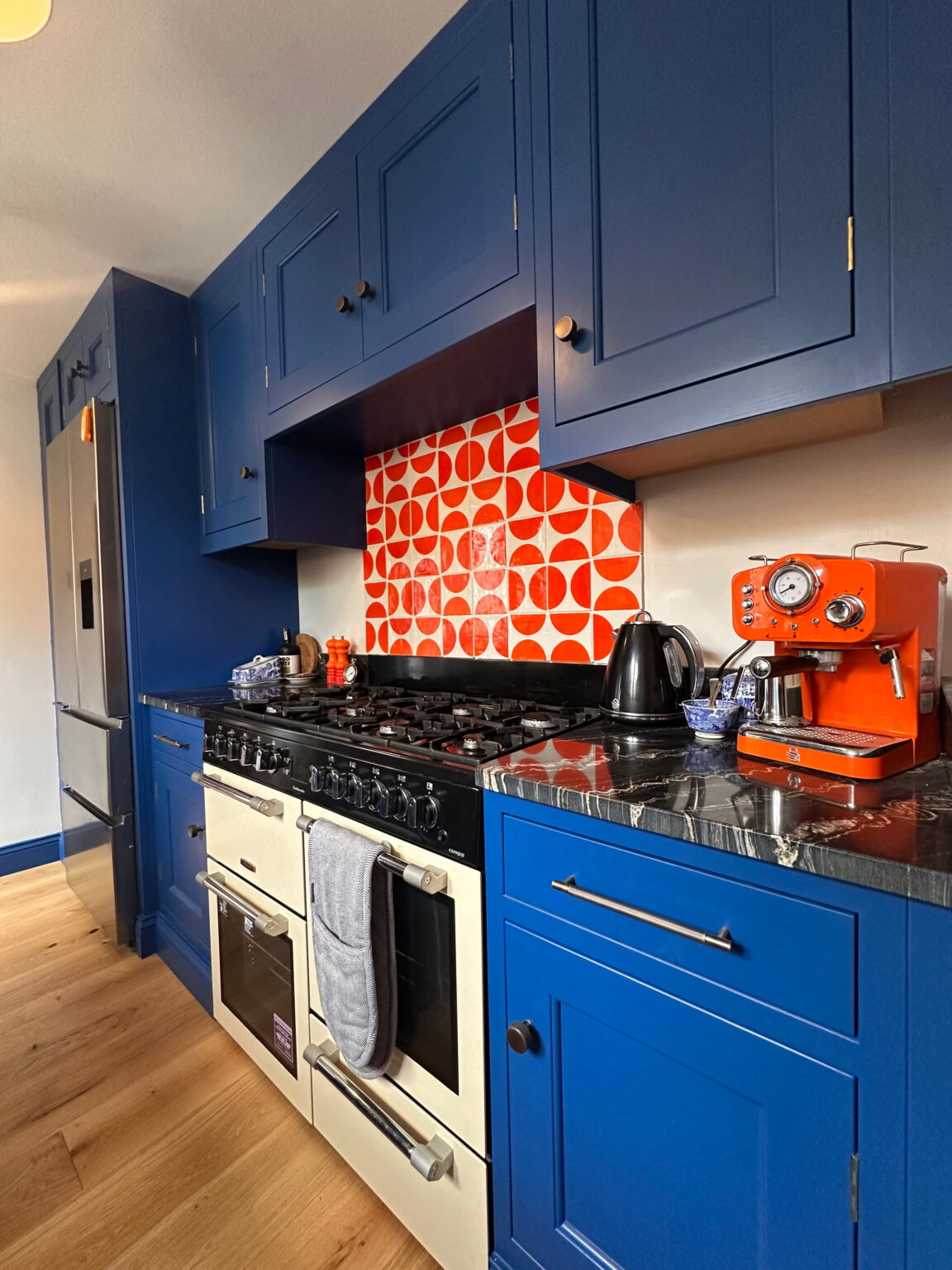 Long Melford Handmade Kitchen by Regent St James 4: Modern blue kitchen featuring a cream range cooker, geometric orange splashback, and black worktop.
