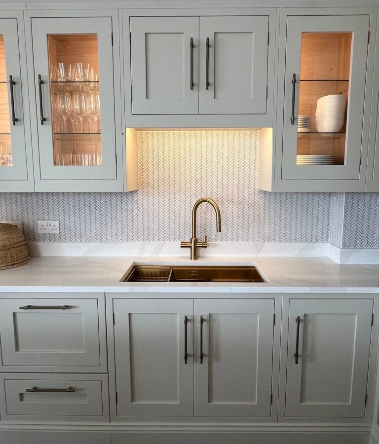 Bespoke kitchen manufacturers: bespoke shaker cabinetry with illuminated glass display cabinets, brass fixtures, and a herringbone tile splashback.